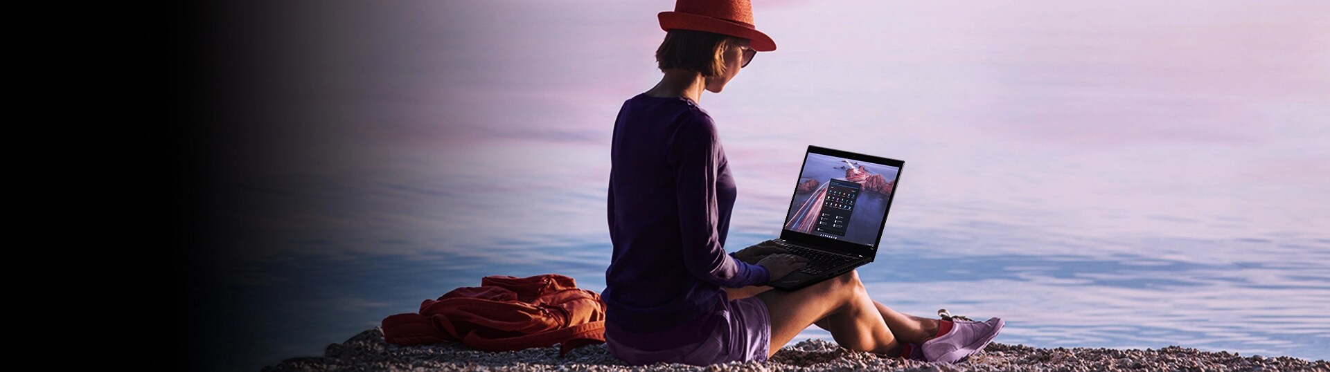 Lady sitting by water with laptop in lap.