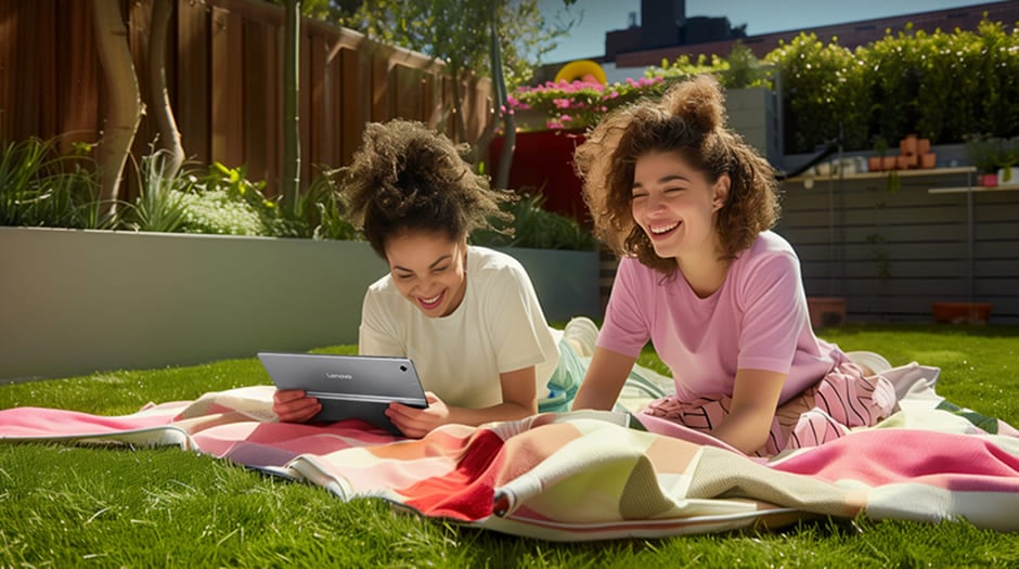 Two young women enjoying a movie outdoors on Lenovo Tab Plus, laughing on a picnic blanket in a sunlit backyard.