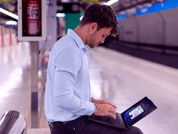 Man sitting on a bench at an empty, clean train station, typing on a Lenovo ThinkPad X13 Gen 4 laptop perched on his lap.