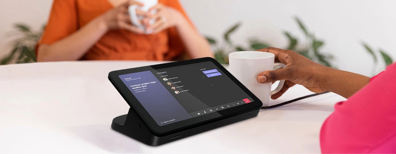 Close up of Lenovo IP Controller, showing Microsoft Teams Rooms interface, on an office desk, next to someone holding a coffee cup, with another person in the background