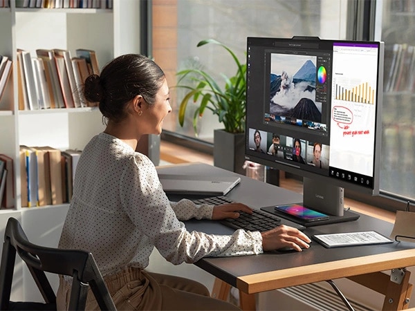 Lenovo ThinkCentre Neo 50a G5 27" all-in-one desktop PC – front right view, showing a woman working on a software using wireless keyboard and mouse, along with having a video conference and the base wirelessly charging a mobile phone in her home office setup.