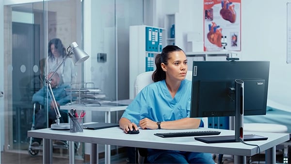 Nurse sat a desk looking at Lenovo ThinkCentre M90a Gen 5 (24″ Intel) all-in-one PC, with patient in wheelchair & another nurse in the background.