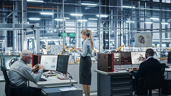 Two engineers in a factory, sat at desks, looking at technical designs on multiple monitors, next to Lenovo ThinkStation P8 workstations, and one colleague standing up, looking at a phone