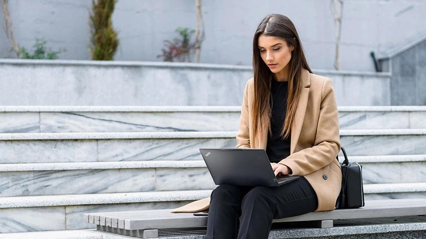 A woman outside sitting on a bench and working on her laptop.
