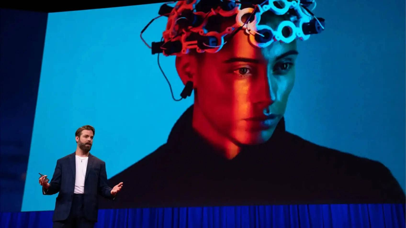 A man presenting on stage with a big image projected behind him of a person in a brain-computer-interfacing wearable device. 