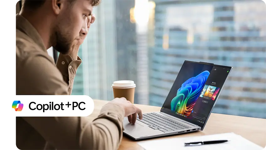 A person working on a Lenovo laptop in a modern office with a cityscape view in the background. The laptop screen shows colorful graphics. A coffee cup and a pen are on the desk beside the laptop. The 'Copilot + PC' logo is displayed in the bottom left corner of the image.