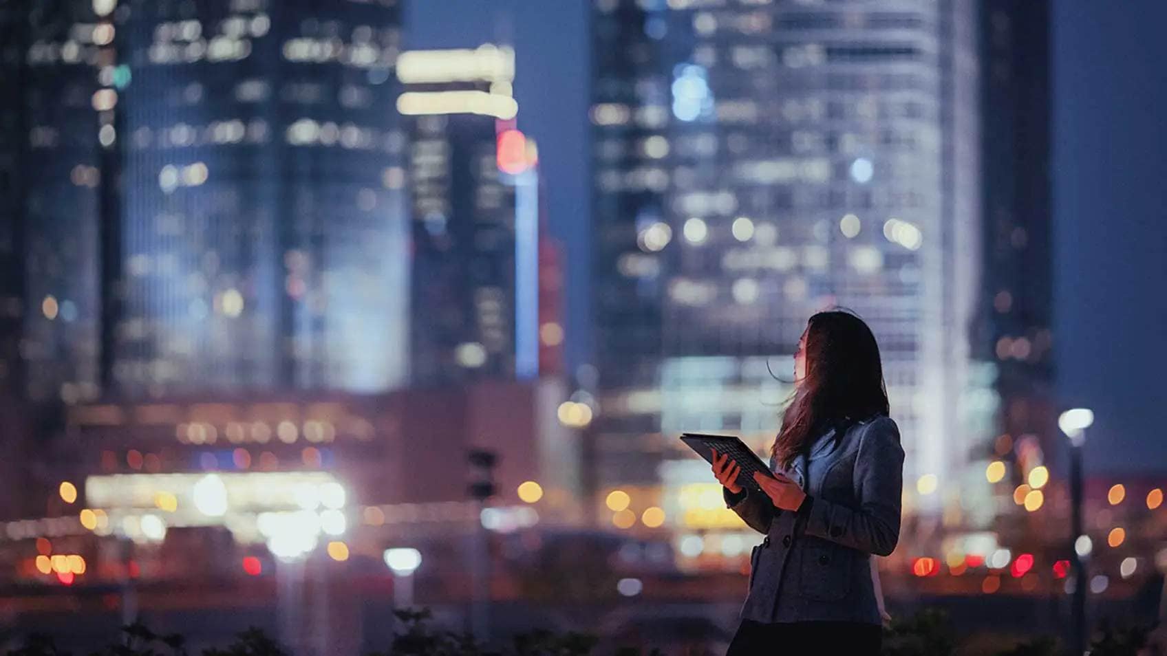 Une femme tenant une tablette et regardant par-dessus son épaule l’horizon de la ville derrière elle, la nuit.