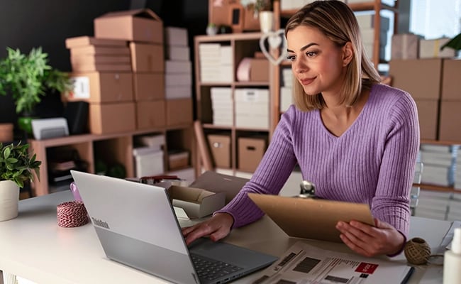 A woman sits at a desk in a storage room holding a clipboard while her other hand controls the Lenovo ThinkBook 16 Gen 9 laptop.