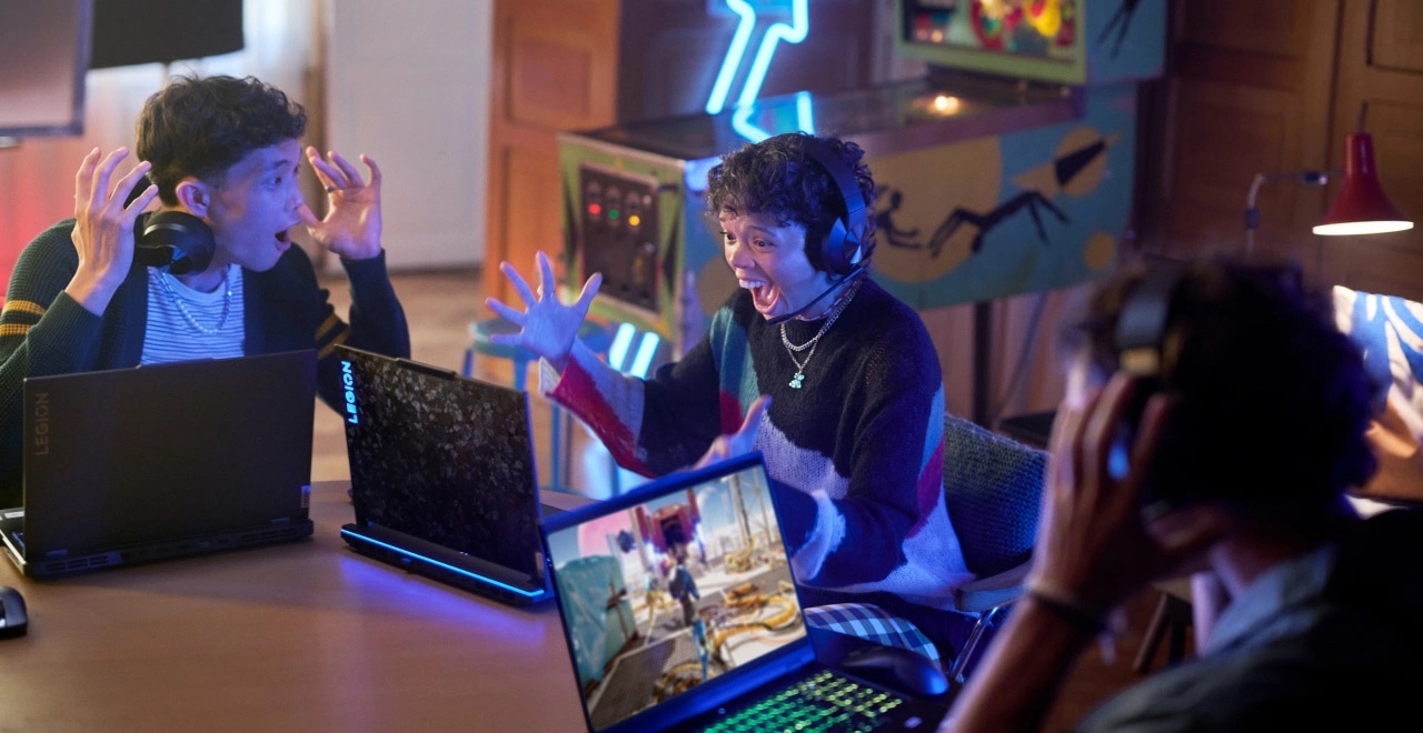 Excited gamers using Lenovo Legion laptops, illuminated by bright screens in a lively gaming room.