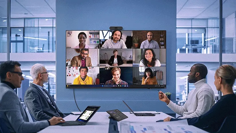 Seis personas sentadas alrededor de una mesa de conferencias en una oficina, participando en una videollamada con otras nueve en una pantalla grande.