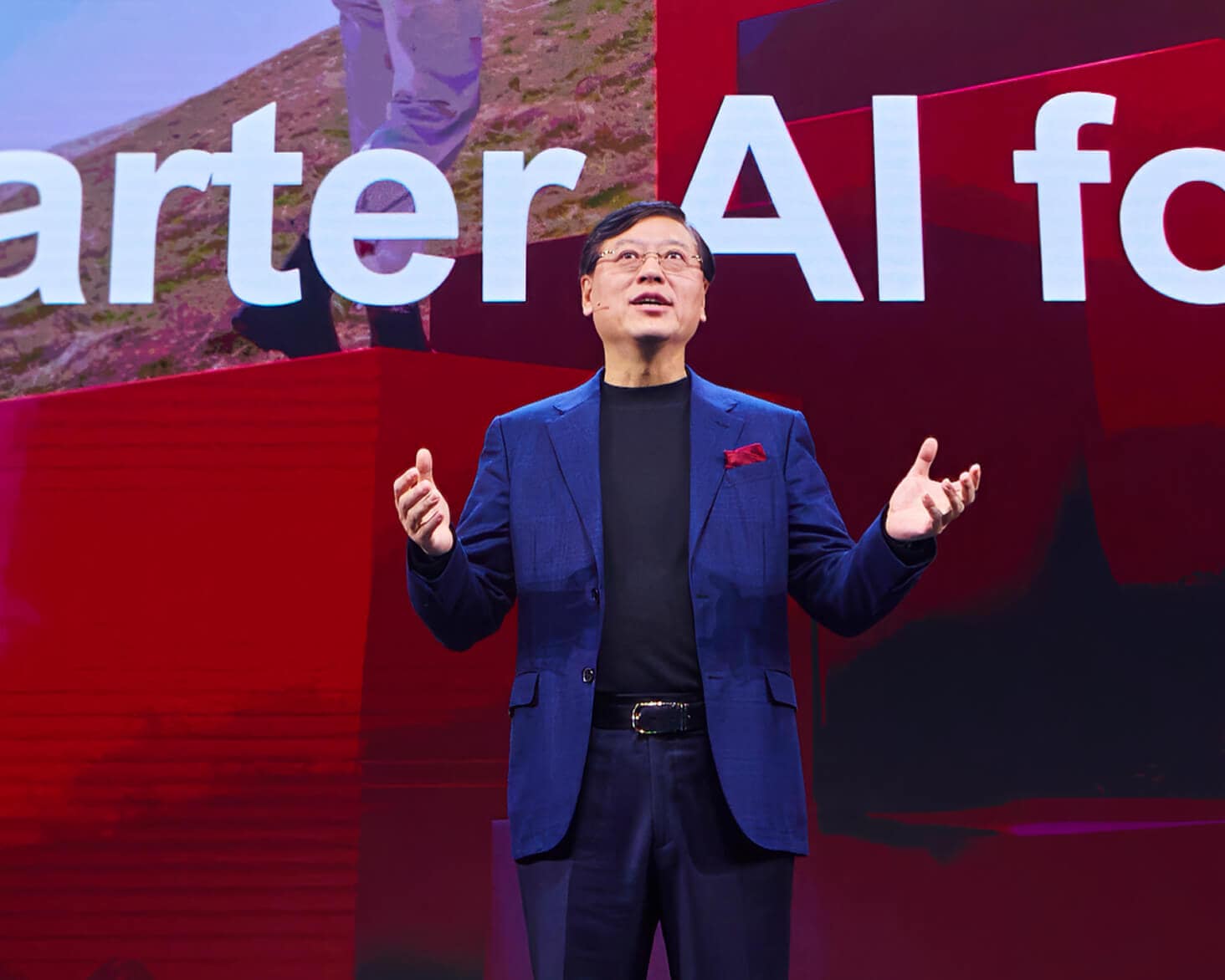 Lenovo's CEO Yuanqing Yang in a blue suit gestures on stage in front of a screen displaying the phrase 'Smarter AI for' with red and black visuals and an outdoor image in the background.