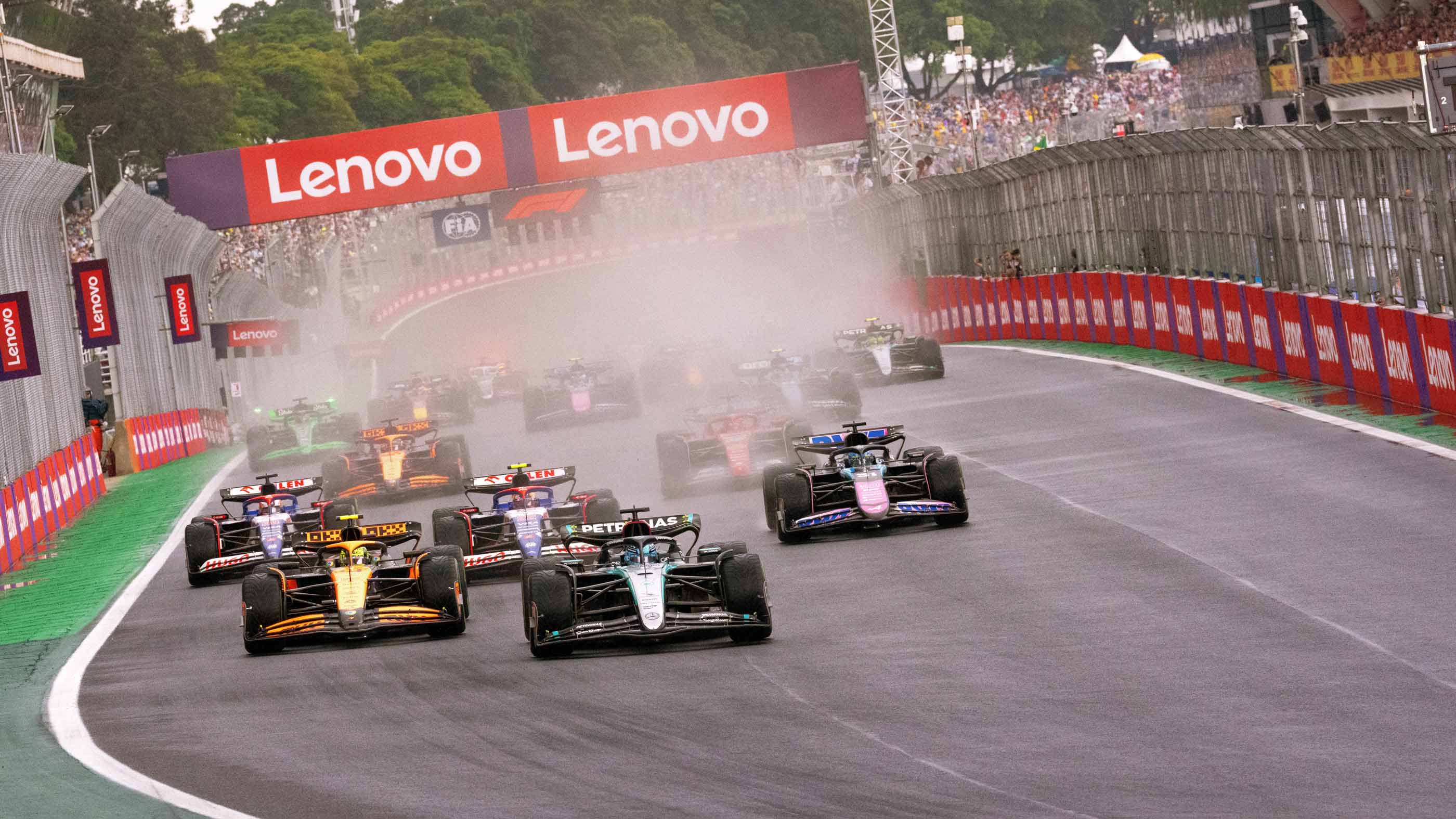 Formula 1 cars racing on a wet track under Lenovo banners, with water spray and crowd in background.