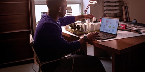 Person sitting at a desk using a laptop while interacting with small architectural or product models, with a desk lamp and organized workspace in a home or office setting.