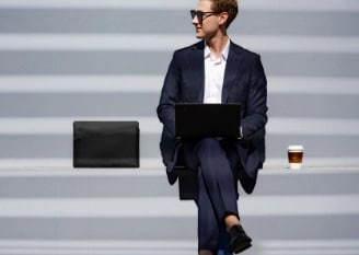 A casually dressed businessman sits on steps alongside his ThinkPad laptop, black laptop carrying case, and a cup of coffee.