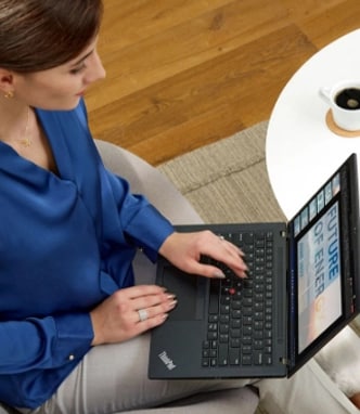 Shot from above, a woman sits at her desk while drinking coffee and shopping online with her Lenovo ThinkPad laptop.