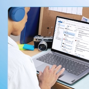 Individual wearing overear blue headphones seated at a desk using a Lenovo laptop displaying the Microsoft Outlook email interface, with a camera, sticky notes and a corkboard in the background
