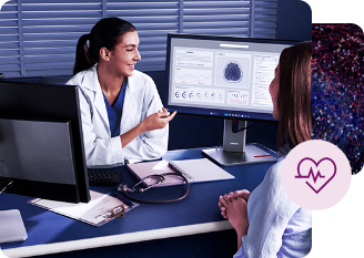 Healthcare professional sitting at a desk with two computer monitors, one displaying medical data and charts, while discussing with a patient in a modern clinical setting.