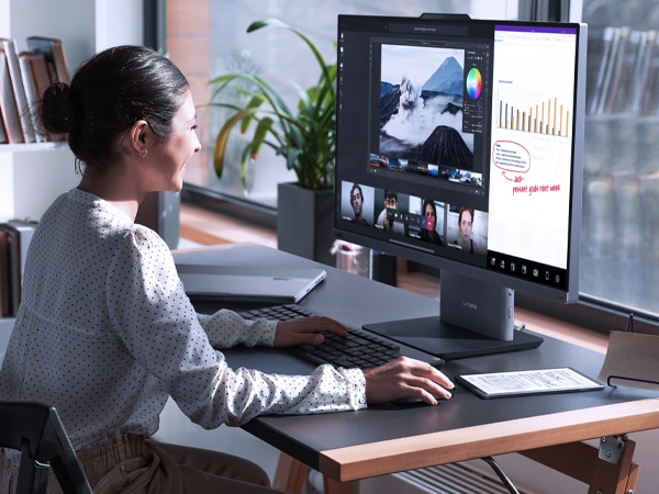 Lenovo ThinkCentre Neo 50a G5 27" all-in-one desktop PC – front right view, showing a woman working on a software using optional wireless keyboard and mouse, along with having a video conference and the base wirelessly charging a mobile phone in her home office setup.