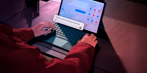 Person using a tablet with a keyboard on a glass table. One screen displays a chat interface, while another features colorful text. The setting is well-lit and has a modern aesthetic