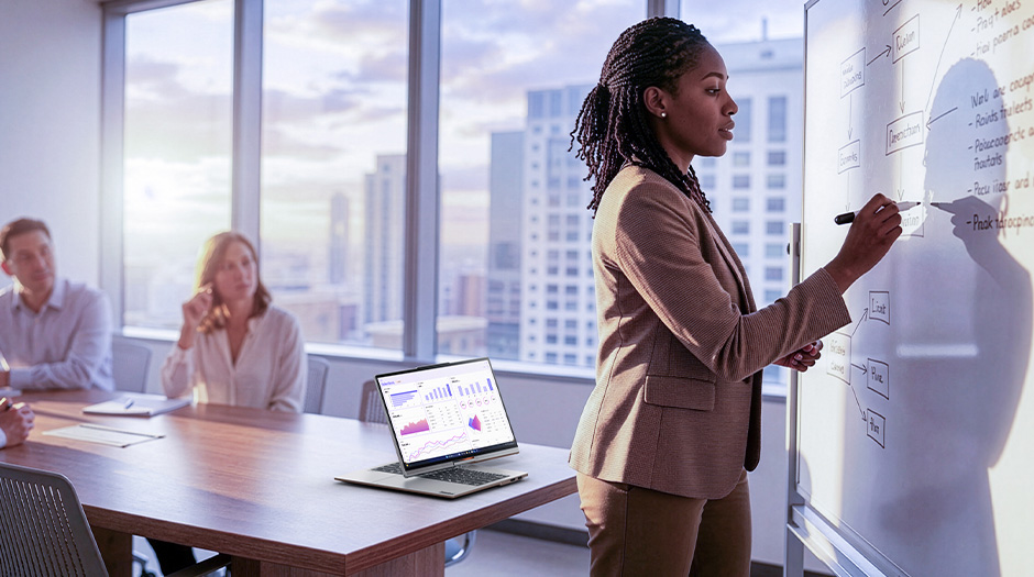 A woman in a meeting room writes on a whiteboard, while a laptop shows charts, highlighting collaboration and presentation features on the Lenovo ThinkBook Plus Gen 7 Auto Twist laptop.