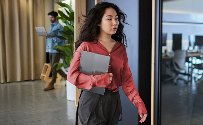 A woman walking in an office carries a lightweight Lenovo ThinkBook Plus Gen 7 Auto Twist (14″ Intel) laptop under her arm.