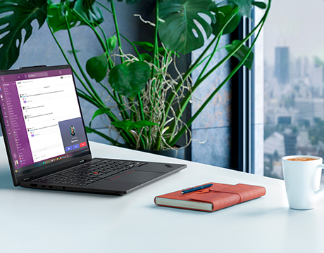 A clean desk with a journal, coffee cup & a Lenovo ThinkPad T14s 2-in-1 Gen 2 (14″ Intel) laptop open, next to a plant & cityscape out the window.