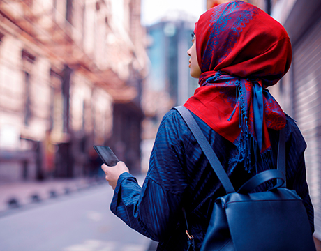 Person in a red scarf walking through an urban street, holding a smartphone.