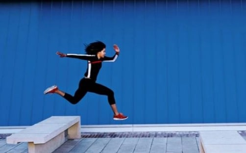 A woman in a tracksuit caught mid-jump over a concrete bench.