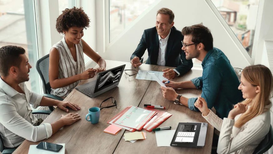 Cinq personnes, trois hommes et deux femmes, assis autour d’une table avec des fournitures de bureau, des verres, une tasse café et 2 portables Lenovo ThinkBook Plus, assis sur la table.