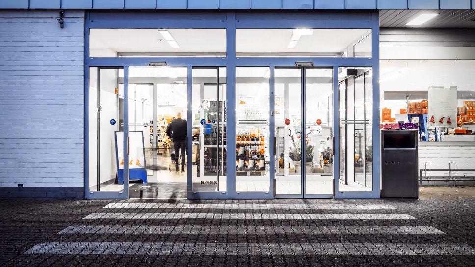 Vue avant d’un magasin de détail bien éclairé avec de grandes portes en verre coulissantes, montrant une personne marchant à l’intérieur.