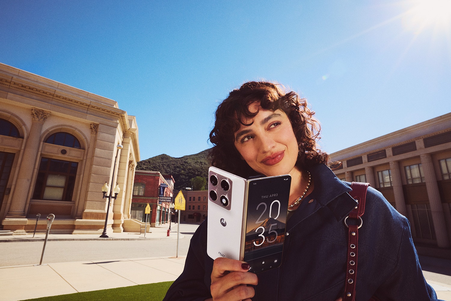 A person holding a Motorola smartphone outdoors with buildings and a sunny blue sky in the background.
