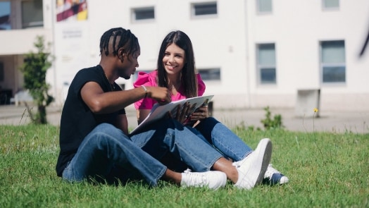 Two coed students are sitting next to each other on a green lawn, working on homework together.