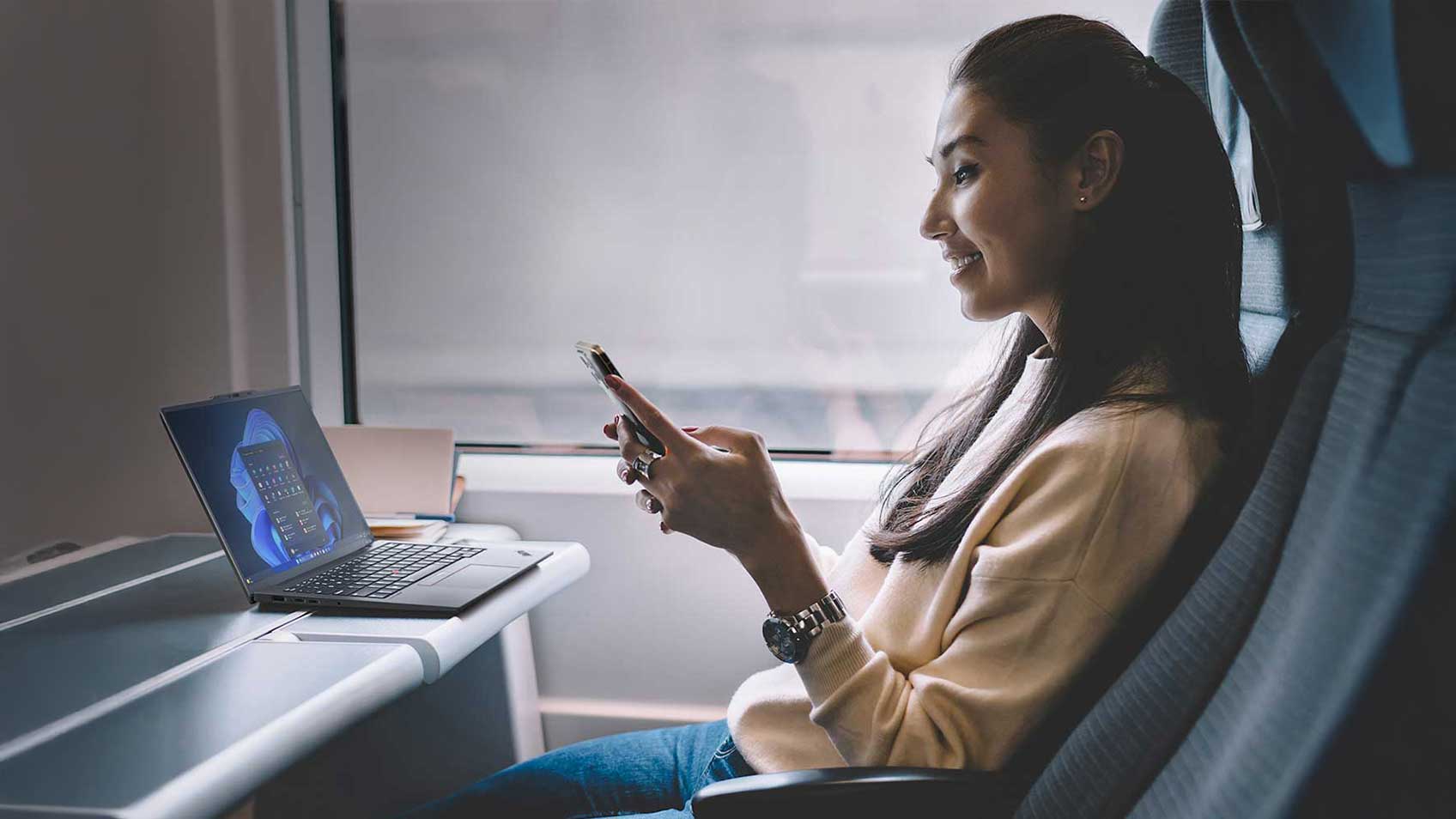 A woman riding on a train and looking at her phone with her laptop on the tray in front of her. 