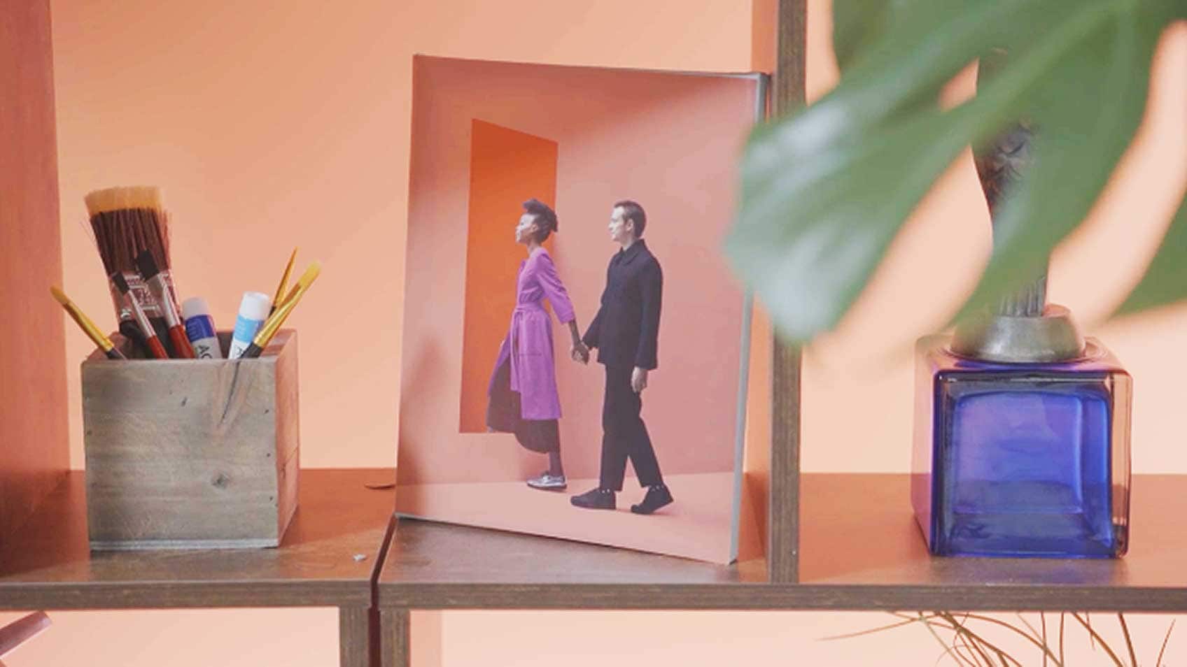 A wooden shelf with a box of paint brushes, a picture of a woman leading a man through the red Lenovo portal, and a plant.