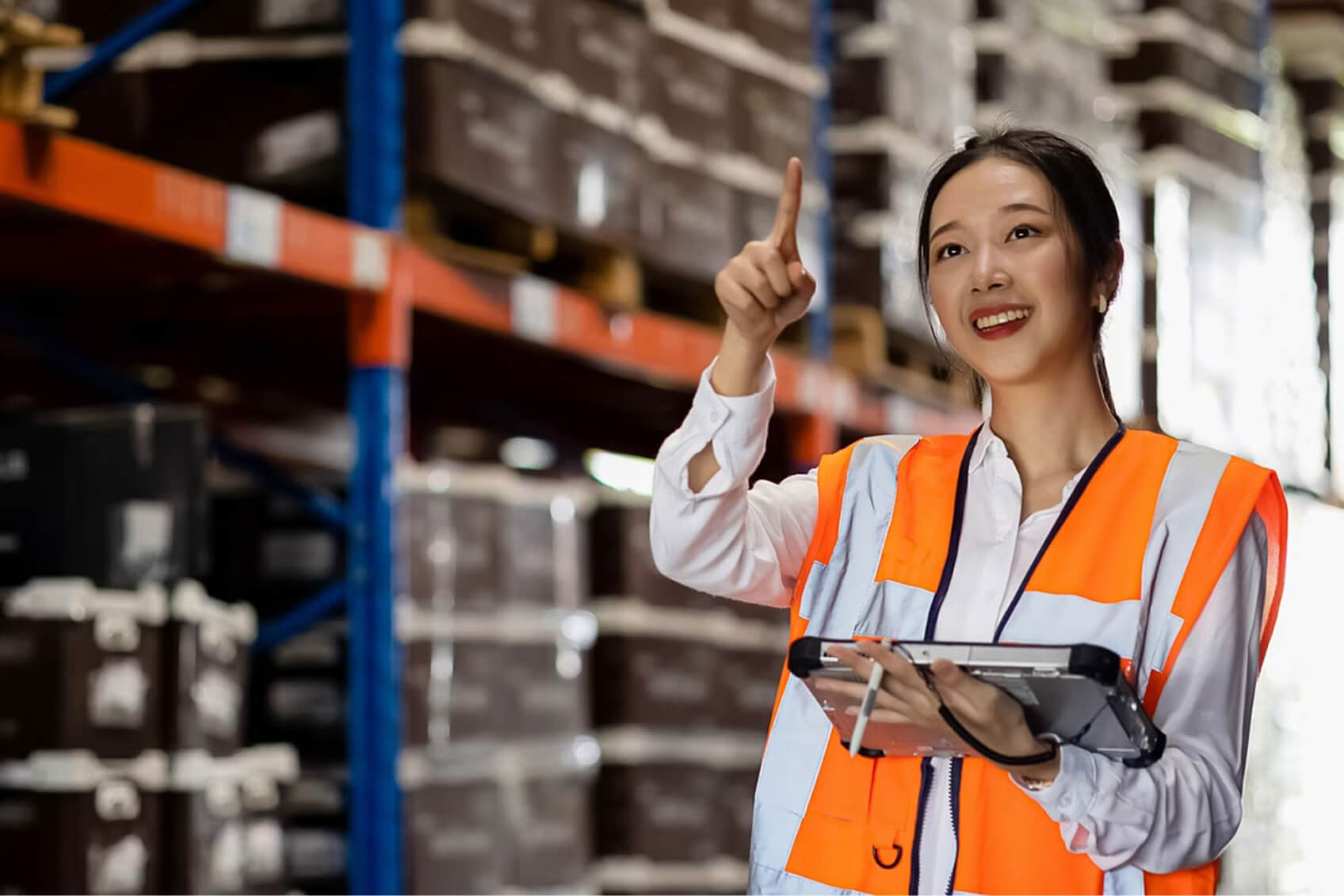 A woman wearing an orange safety vest, holding a tablet, and pointing at a shelf in a warehouse. 
