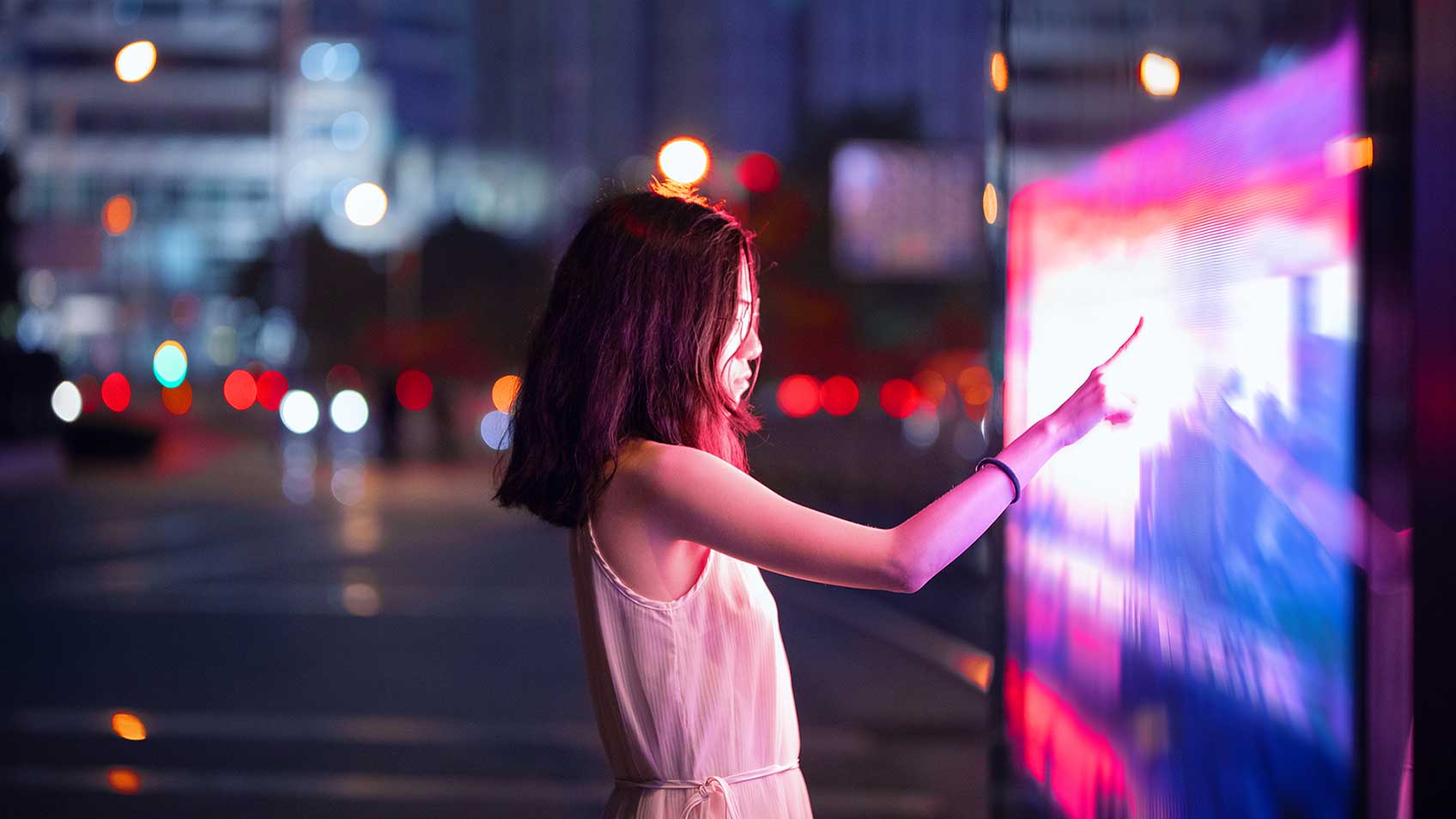 Une fille touchant un panneau interactif et illuminé la nuit en ville.