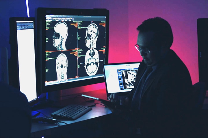A large monitor displaying brain scans and a person analyzing on a tablet in a dimly lit medical setting.
