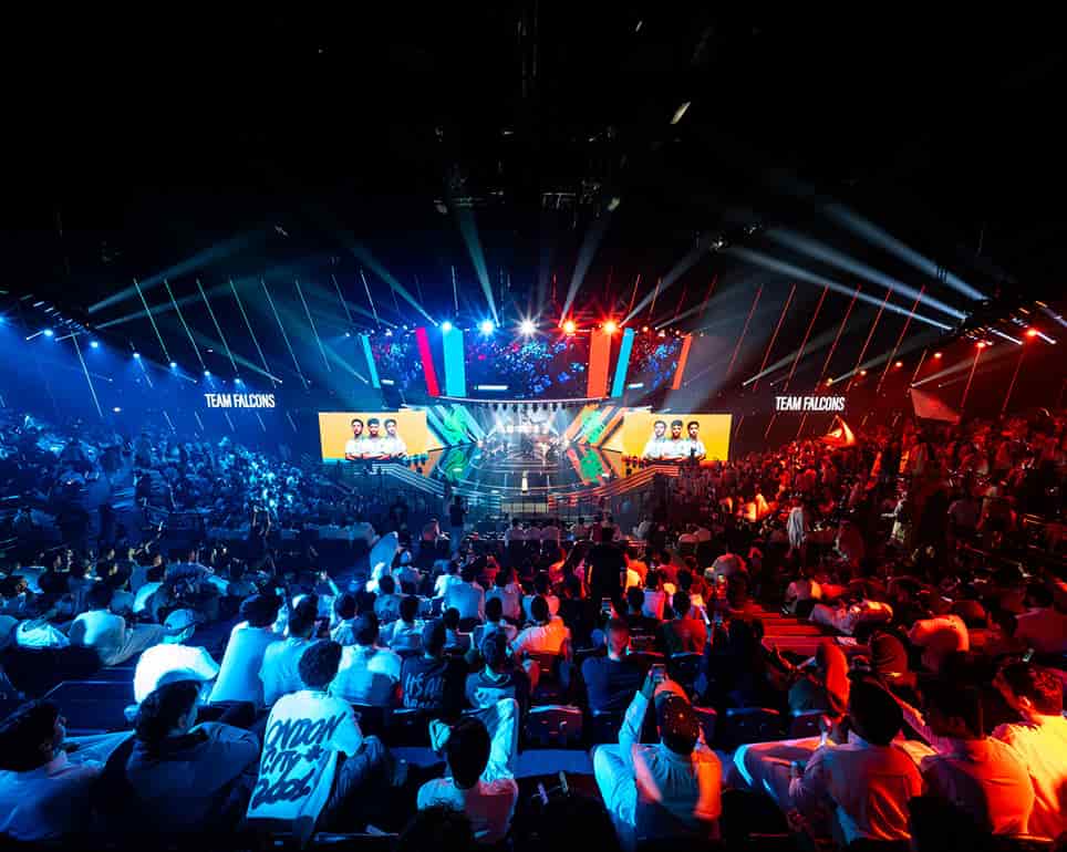 Vista amplia de la arena de esports de EWC llena de aficionados, iluminación del escenario dramática y el evento mostrado en pantallas masivas.