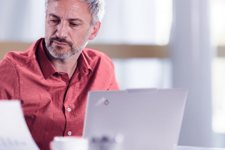 Business professional reviewing documents while working on a Lenovo ThinkPad in a bright office.